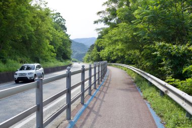 Gongju, Güney Kore - 27 Mayıs 2021: Çevresi yemyeşil ağaçlar ve yuvarlanan tepelerle çevrili, bisikletçiler ve gezginler için manzaralı bir rota sunan, kendini adamış bir bisiklet yolu kırsal bir yola paralel gider..