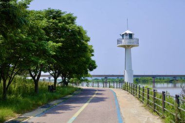 Nonsan, Güney Kore - 27 Mayıs 2021: Ağaçlarla kaplı manzaralı bisiklet yolu Ganggyeong yakınlarındaki Geum Nehri 'ne bakan beyaz bir gözetleme kulesine çıkıyor. Sakin nehir kenarı manzarası bisiklet ve yürüyüş için huzurlu bir yer sunuyor..