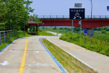 Iksan Şehri, Güney Kore - 28 Mayıs 2021: Kırmızı köprünün altından dönen bir bisiklet yolu geçiyor, kaygan bir bölge hakkında bisikletçileri uyaran bir uyarı işareti var. Bir çardak yol boyunca dinlenme yeri sağlar..