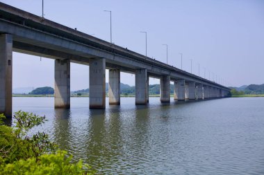 Gunsan, South Korea - May 28, 2021: The Geum River Grand Bridge extends across the calm waters of the Geum River, supported by towering concrete pillars, with rolling hills and greenery in the background. clipart