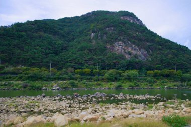 Önünde kayalık bir nehir kıyısı olan Seomjin Nehri üzerinde engebeli, ormanlı bir dağ yükseliyor. Manzara bölgeleri vurgular. Doğal güzellik ve jeolojik çeşitlilik.