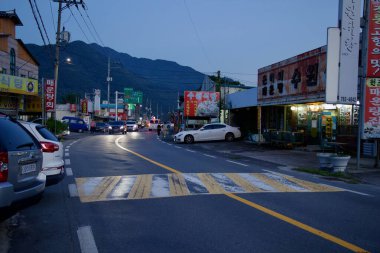 Gurye İlçesi, Güney Kore - 3 Ekim 2021: Küçük bir kasabada hareketli bir cadde alacakaranlıkta canlanıyor, yerel restoranlar, park edilmiş arabalar ve yol kenarlarını bir dağ zeminine doğru aydınlatan vitrin tabelaları var..