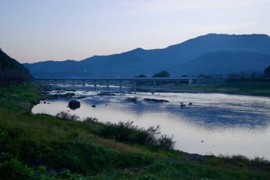 Gurye İlçesi, Güney Kore - 3 Ekim 2021: Seomjin Nehri alacakaranlıkta sakin bir şekilde bir köprünün altından akar, gün batımını yansıtır. Çevresindeki dağlar ve nehir kıyıları huzurlu ve resimsel bir manzara yaratıyor..