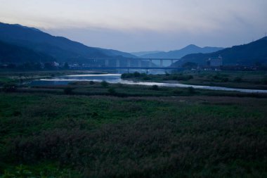 Gurye İlçesi, Güney Kore - 3 Ekim 2021: Seomjin Nehri alacakaranlıkta vadiden süzülen gökyüzünü yansıtıyor. Uzak köprüler ve küçük köyler, yuvarlanan dağların arasına yerleşmiş, huzurlu bir kırsal alan yaratıyor..