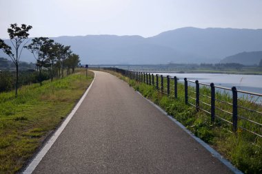 Gurye County, Güney Kore - 3 Ekim 2021: Nehir boyunca uzanan sakin ve iyi döşenmiş bir bisiklet yolu, uzak dağlar ve puslu bir öğleden sonra gökyüzü manzaralı manzaralı bir rota sunuyor..