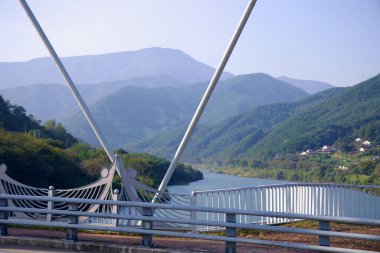 Gurye County, Güney Kore - 3 Ekim 2021: Namdo Köprüsü 'nden görülen sisli bir dağ perdesi Seomjin Nehri' ni kareler. Köprünün kabloları ve süslü tırabzan kontrastı yemyeşil ve uzak yamaçlardaki evlerle.