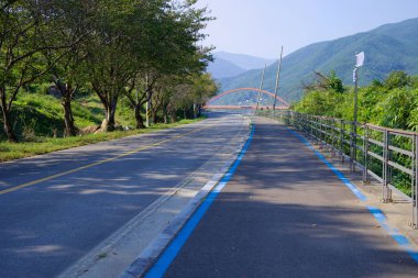 Gurye İlçesi, Güney Kore - 3 Ekim 2021 Seomjingang Bisiklet Yolu 'nun manzaralı bir uzantısı açık bir sonbahar gününde yemyeşil ve dağ manzarasıyla çevrili kırmızı kemer köprüye doğru esiyor..