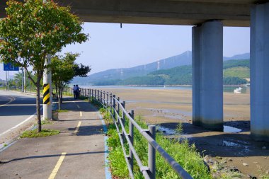 Gwangyang, Güney Kore - 3 Ekim 2021 Taein Adası 'nı çevreleyen yol boyunca uzanan manzaralı bir bisiklet ve yaya yolu, Seomjin Nehri' nin gelgit düzlüklerini ve uzak dağlarını gören bir köprünün altından geçer..