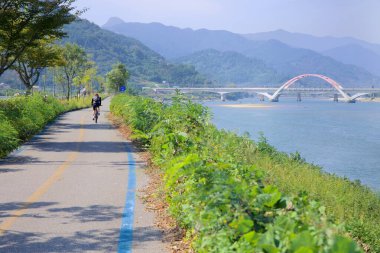Gwangyang, Güney Kore - 3 Ekim 2021 Seomjingang Bisiklet Yolu boyunca bir bisikletçi, Seomjin Nehri 'ne paralel gidiyor. Arka planda, modern kemerli bir köprü, yemyeşil dağların ortasındaki nehri kaplıyor..