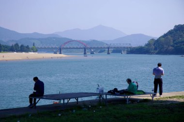 Gwangyang, Güney Kore - 3 Ekim 2021: İnsanlar Seomjin Nehri boyunca oturur ve dinlenirler, kırmızı kemerli demiryolu köprüsü ile sisli dağların arkaplanına doğru uzanır..