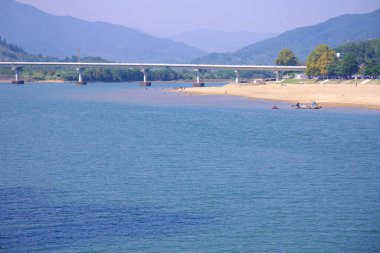 Gwangyang, Güney Kore - 3 Ekim 2021 Seomjin Nehri manzaralı bir manzara, burada bir kumsal ziyaretçileri suyun keyfini çıkarıyor. Nehir boyunca uzanan modern bir köprü, dağlık araziyi birbirine bağlıyor..