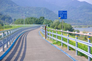 Gwangyang, Güney Kore - 3 Ekim 2021 Seomjingang Bisiklet Yolu eğrileri boyunca pürüzsüz ve bakımlı bir bölüm, güvenlik korkulukları ve manzaralı dağ manzarası boyunca bisikletçilere rehberlik eden yön tabelaları.