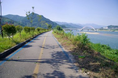 Gwangyang, Güney Kore - 3 Ekim 2021 Seomjingang Bisiklet Yolu 'nun manzaralı bir uzantısı Seomjin Nehri' ni takip ederek uzak dağları olan modern kemerli bir demiryolu köprüsüne doğru ilerliyor..