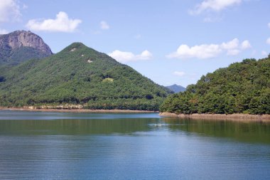 Damyang İlçesi, Güney Kore - 23 Eylül 2020: Damyang Gölü 'nün durgun suları çevresindeki orman tepelerini ve dağları yansıtıyor, berrak bir gökyüzü altında barışçıl ve manzaralı doğal bir manzara yaratıyor..