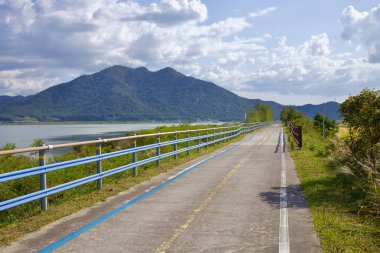 Muan County, Güney Kore - 25 Eylül 2020: Yeongsangang Bisiklet Yolu nehre paralel akar, mavi güvenlik korkulukları ve yemyeşil ile sınırlıdır, parlak bir gökyüzünün altında yükselen bir dağ sırası vardır..