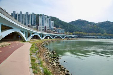 Ulsan City, Güney Kore - 31 Mart 2025: Kırmızı bir bisiklet yolu ve yaya geçidi Taehwa Nehri 'nin kıvrımlarını sessiz bir yerleşim bölgesi yakınlarındaki bir dizi beyaz kemerli köprünün altında takip ediyor..