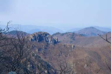 Ulsan City, Güney Kore - 2 Nisan 2025 Yeongnam Alpleri 'nin yamaçlarından dramatik bir kayalık oluşumu, kurak bahar ormanları ve puslu bir gökyüzü altında uzak tepelerle çevrili..