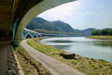 Ulsan City, Güney Kore - 31 Mart 2025: Taehwa Nehri boyunca uzanan bir köprünün kemerli desteklerinin altından geçen nehir kenarı yürüyüş yolu, Simnidae Ormanı 'nı yavaşça geçerek eteklerin ötesine doğru kıvrılıyor..