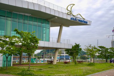 Incheon, Güney Kore - 19 Mayıs 2025: Arka planda Ara Waterway boyunca yelkenlilerin limana yanaştığı Ara Marina 'daki cam duvarlı çadırı çevreleyen ağaçlı yeşil bir çim.