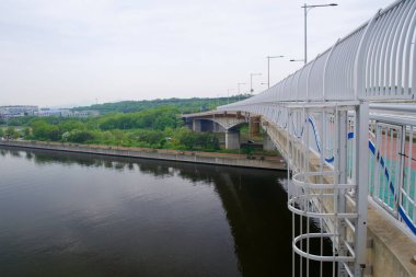 Incheon, Güney Kore - 19 Mayıs 2025: Cheongungyo Köprüsü kavisli bir yaya ve bisiklet yolu ile Ara Nehri boyunca uzanır ve aşağıdaki Ara Batı Deniz Kilidi 'nin kuzey girişinde ağaçlandırılmış bir kanal yolu ve sakin bir su vardır..