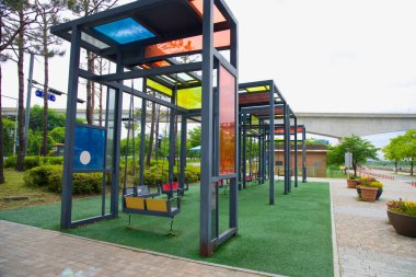 Incheon, Güney Kore - 19 Mayıs 2025: Renkli cam ve çelikten yapılmış modern bir salıncak Sicheon Waterside Park 'taki Ara Waterway yakınlarındaki çam ağaçlarının altında tasarımla harmanlanmış bir oyun..
