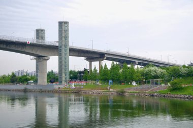 Incheon, Güney Kore - 19 Mayıs 2025: Ara Waterway 'den Gyeyang Köprüsü' ne kadar uzanan iki cam asansör kulesi, manzaralı bir parka ve renkli heykeller ve bahar ağaçlarıyla nehir kenarındaki yola bakıyor..