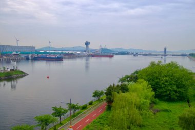 Incheon, Güney Kore - 19 Mayıs 2025: Batı Denizi Kilidi yakınlarındaki Ara Waterway ve Incheon Limanı 'nın yemyeşil ve sanayi kıyılarına paralel uzanan kırmızı yaya geçidi tarafından çerçevelenmiş panoramik manzarası.