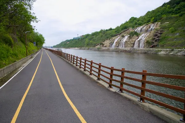 Incheon, Güney Kore - 19 Mayıs 2025: Ara Şelalesi Gyeongin Ara Su Yolu boyunca teraslı bir kayalıktan aşağı akarken Ara Bisiklet Yolu bulutlu bir yay altında sakin bir kanalın yanında devam ediyor..