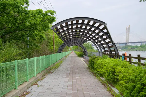 Incheon, Güney Kore - 19 Mayıs 2025: Ara Su Yolu 'nun yanındaki Ara Bisiklet Yolu' nun bir kısmını kaplayan modern kemerli gölgelik, Incheon Köprüsü 'nü ilkbahar yeşilliği ve asfalt dinlenme alanının ötesindeki mesafede görebilirsiniz..