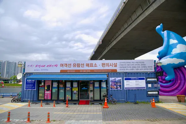 Incheon, Güney Kore - 19 Mayıs 2025: Ara Cruise hizmetleri için Sicheon Rıhtımı bilet gişesi Sicheon Köprüsü 'nün altında, Sicheon Waterside Parkı boyunca uzanan Sicheon Waterside Park' taki canlı Bisikletli Bulut Adam heykelinin yanında duruyor..