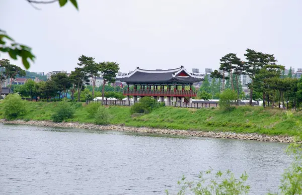 Incheon, Güney Kore - 19 Mayıs 2025: Suhyangwon Pavyonu Ara Waterway 'deki nehir kıyısının üzerinde yükseliyor, yüksek geleneksel tasarımı çam ağaçları ve şehir binaları tarafından çerçeveleniyor..