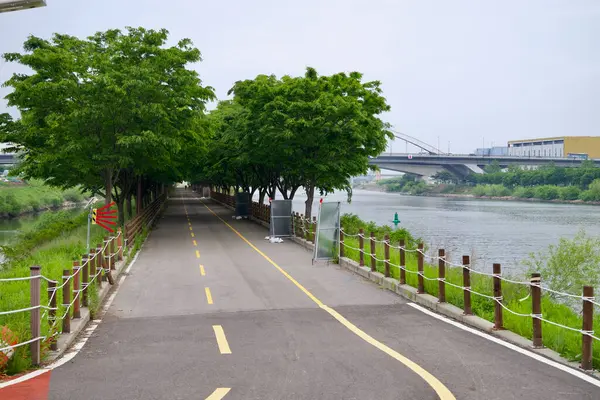 Incheon, Güney Kore - 19 Mayıs 2025: Ara Bisiklet Yolu, Ara Su Yolu boyunca ağaçlardan oluşan yoğun bir gölgelik altında devam ederken, arka planda Ara Batı Deniz Kilidi köprüsü görülebiliyor..