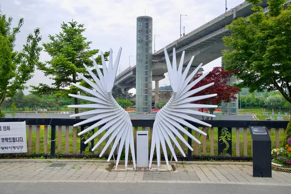 Incheon, Güney Kore - 19 Mayıs 2025: Melek kanadı şeklinde beyaz metal bir heykel, arkasındaki Gyeyang Köprüsü asansör kulesini çerçeveler ve Ara Waterway parkı boyunca popüler bir fotoğraf noktası sunar..