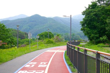 Namyangju City, South Korea - May 19th 2025: Curving red-asphalt bike lane beside a disused railway and Bukhan River valley mountains on the Gyeongchun Line greenway invites cyclists and walkers.