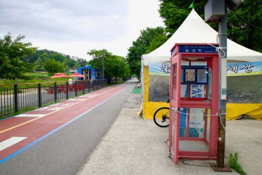 Neungnae İstasyonu Sertifika Merkezini gösteren kırmızı bir kabin Bukhangang Bisiklet Yolu 'nun yanında duruyor, tren kafesi ve gölgeli dinlenme alanları uzaktan görülebiliyor..