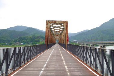 Yangpyeong County, South Korea - May 19th 2025: The rusted lattice of the Bukhangang Railway Bridge rises over the river, with a clear view of Yangsu Station and nearby hills under a pale overcast sky.