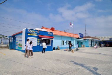 Gangeung Şehri, Güney Kore - 1 Haziran 2025: Turuncu çatılı ve ulusal bayraklı gök mavisi Jeongdongjin İstasyonu binasının önünde parçalı bulutlu bir sabah gökyüzü altında ziyaretçiler duruyor.