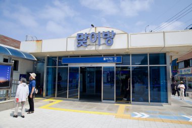 Gangeung Şehri, Güney Kore - 1 Haziran 2025: Yolcular Jeongdongjin İstasyonu 'nun peron girişine yakın yürüyorlar, parlak bir gökyüzü altında mavi cam kapılar ve yön işaretleriyle işaretlenmişler..