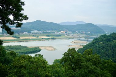 Gongju, Güney Kore - 13 Haziran 2025: Sarmal Geum Nehri, kum barları, orman tepeleri ve bulutlu bir gökyüzü altındaki şehir manzarasına bakan Gong Dağı Kalesi 'nden yükseltilmiş bir manzara.