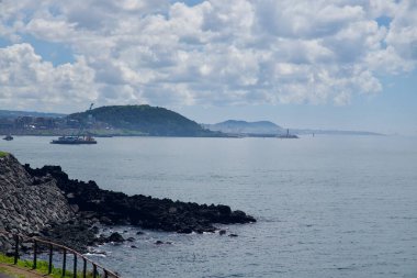 Koyu lav kayaları Eoyeong Park 'tan yeşil bir buruna, çalışan mavnalara ve yükselen bulutların altındaki liman dalgakıranlarında kırmızı deniz fenerlerine doğru bir manzara oluşturur..