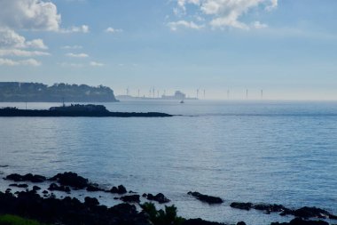 Aewol sahili boyunca puslu bir manzara. Açık deniz rüzgar çiftliği ve ufukta sanayi kompleksi, balıkçı dalgaları ve ön planda karanlık lav kayaları var..