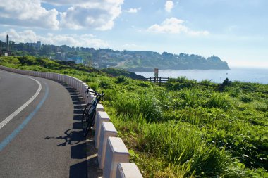 Bir yol bisikleti Hagu-Aewol Sahil Yolu boyunca alçak bloklara yaslanıyor, mavi benekli ve yeşil burunlu sahil kasabasına doğru koşuyor..