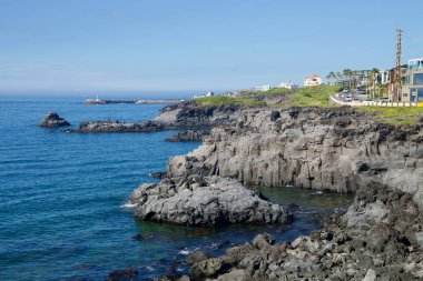 Katmanlı bazalt kayalıkları Gueom kıyısındaki dar körfezleri çevreleyen liman girişi ve deniz feneri berrak mavi gökyüzünün altında görülebilir..