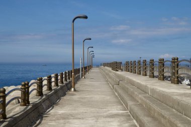 Iho Limanı 'ndaki dalgakıran boyunca simetrik geçitte denize açılan bronz korkuluklar ve sokak lambaları, altında tetrapod zırhı ve yukarıda mavi gökyüzü bulunur..