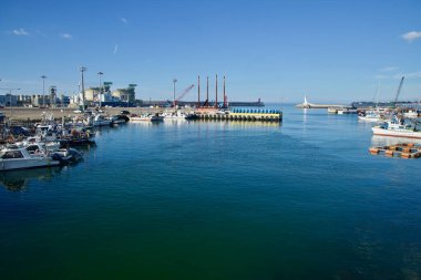 Aewol Limanı 'nda geniş görüş alanı, açık bir yaz gökyüzü altında liman ağzını koruyan beyaz ve kırmızı deniz fenerleri ile solda demirlemiş balıkçı tekneleri, vinçler ve KOGAS LNG tanklarını gösteriyor..
