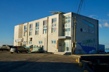 Jeju Hansupul Haenyeo Okulu 'nun beton binası rıhtımda duruyor. Tabelaları ve balina tabelaları limana bakıyor. Yakındaki balıkçılık operasyonlarından kalma araçların ve teçhizatların yanında..