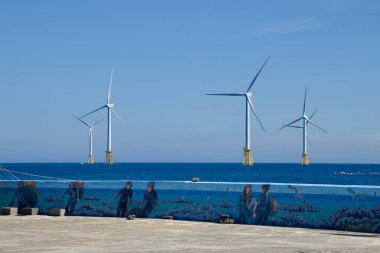 Jeju Haenyeo dalgıçlarıyla boyanmış liman duvarı denize bakıyor ve ufukta mavi suların ötesinde dönen rüzgar türbinleri var..