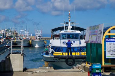 Biyangdo yolcu feribotu Hanlim Limanı 'nda manevra yapıyor. Bir güverte hazırda bekliyor ve yakınlardaki tabelalar yelken açıyor. Sıralar halinde kalamar balıkçı tekneleri dalgakıran boyunca demirliyor..