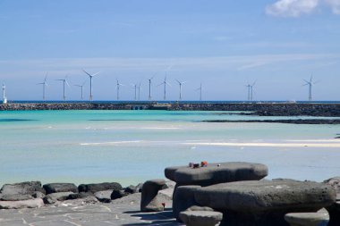 Hyeopjae gezinti güvertesinde bazalt piknik masaları türkuaz sığlıkları gözden kaçırır ve açık deniz rüzgar türbinleri ve beyaz bir deniz feneri ile kaplı bir dalgakıran,.