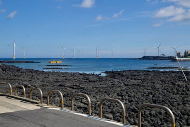 Açık deniz rüzgar türbinleri, Jeju Adası 'ndaki Hanlim yakınlarında bir bazalt kıyı şeridinin ötesinde döner. Sığ gel-git havuzları ve dağınık bulutların altında sakin mavi Kore Boğazı' nı çevreleyen düşük dalgalı bir su tabakası vardır..